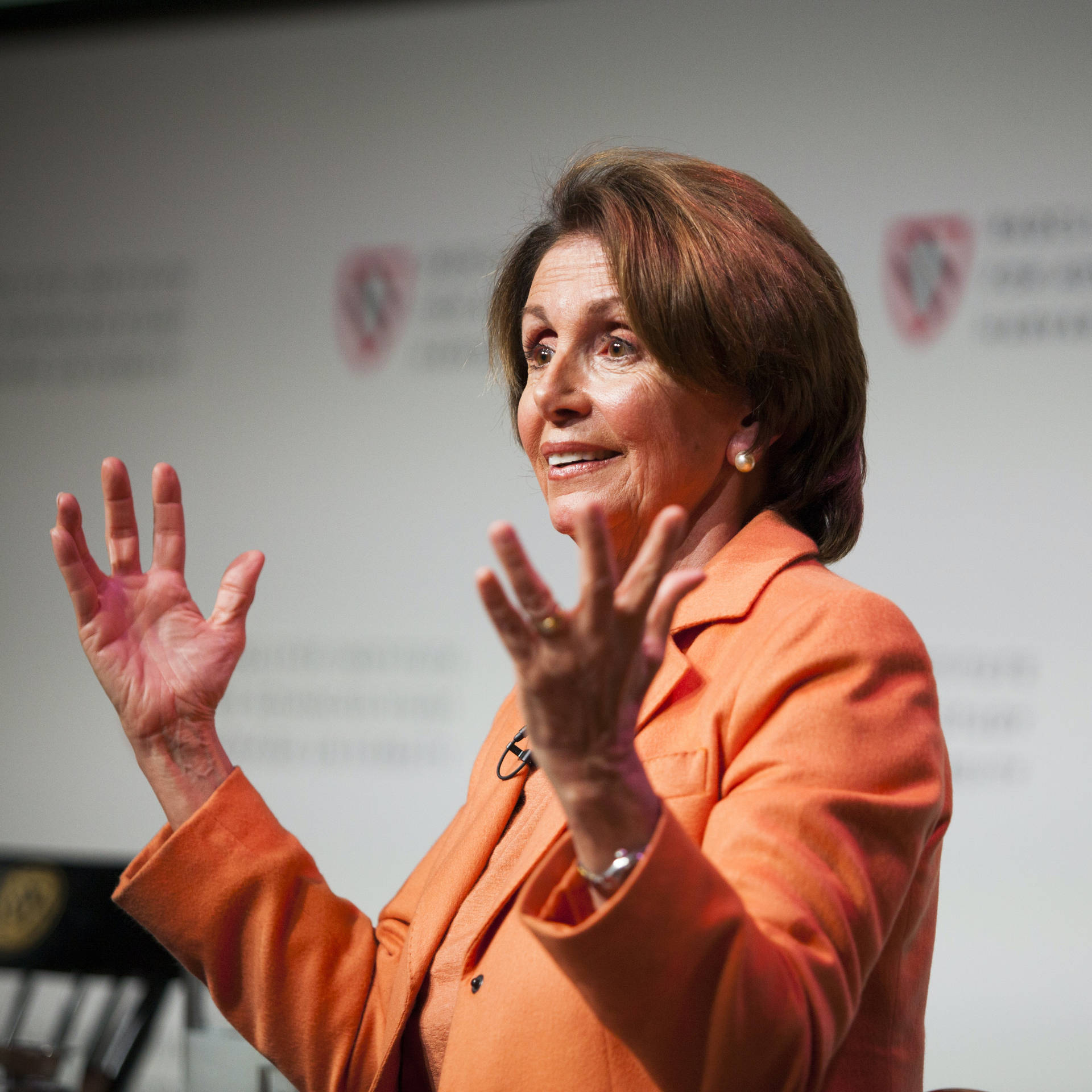 Nancy Pelosi's Bold Presence In Orange Attire Background