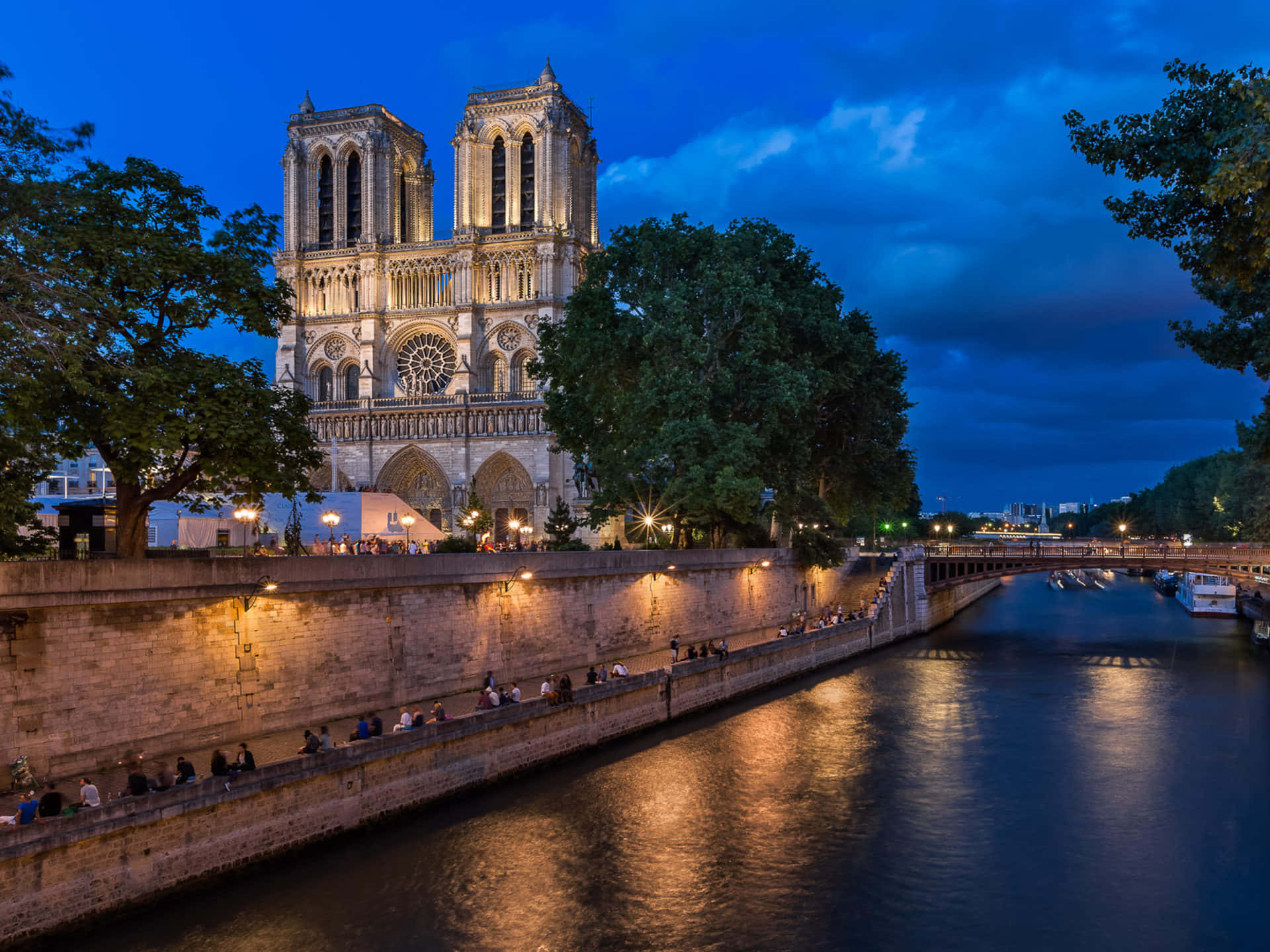 Notre Dame Cathedral With Tourists On The River Pathway Background