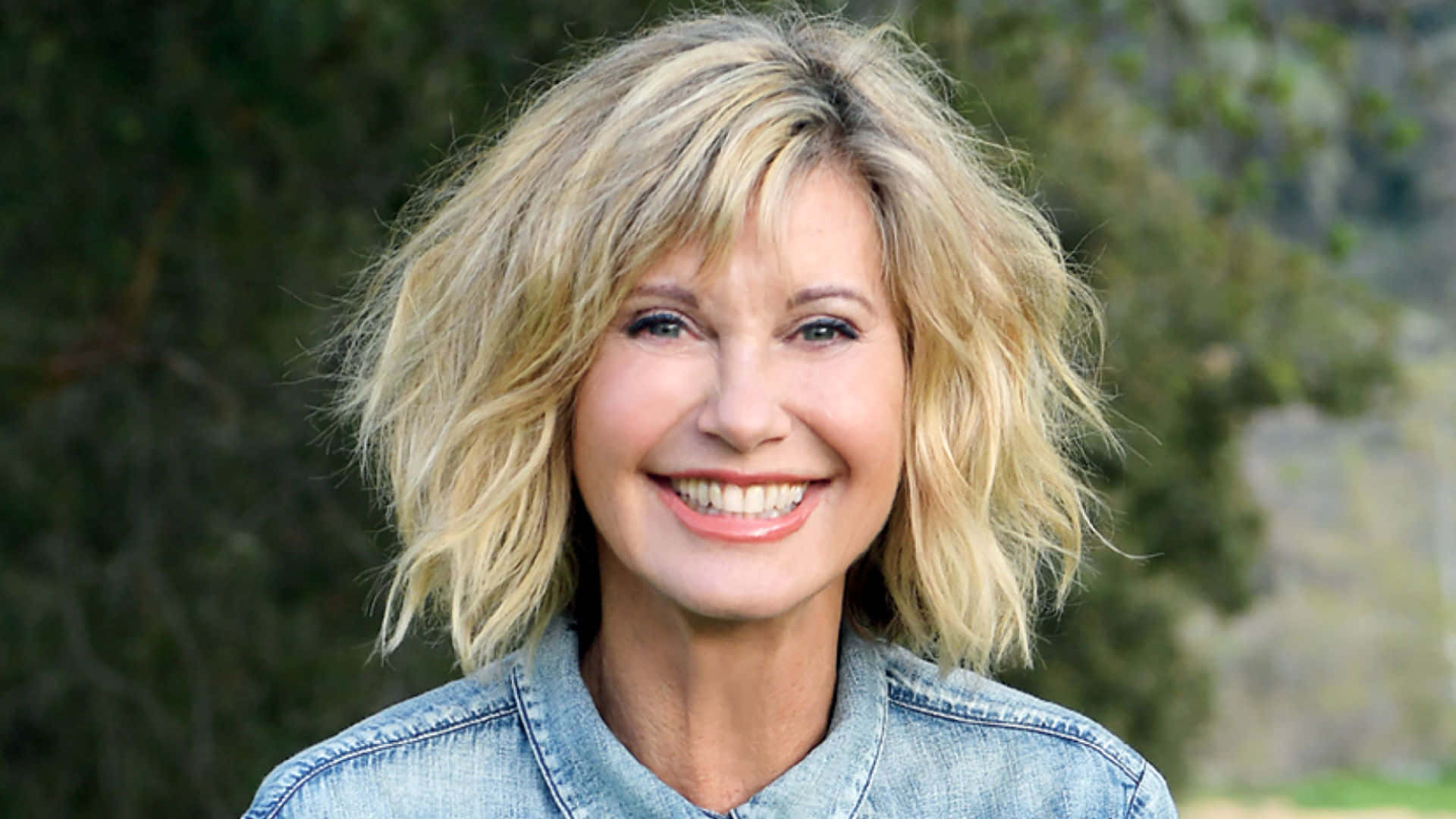 Olivia Newton John Against Blurry Green Trees Background