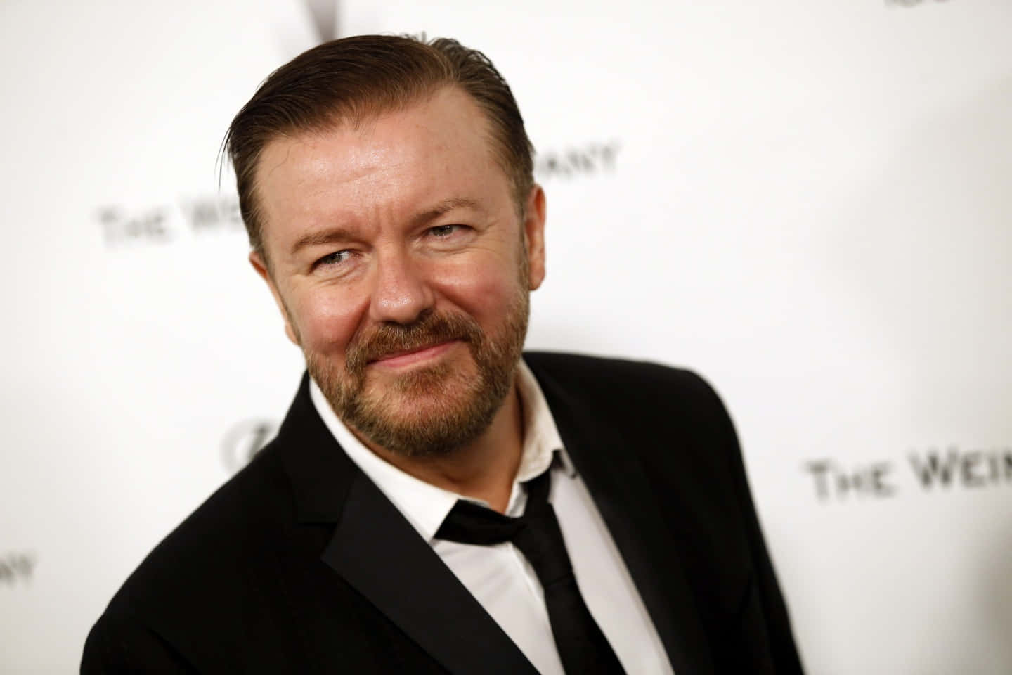 Ricky Gervais, Adorned With A Smart Casual Look, Smiling At The Camera. Background