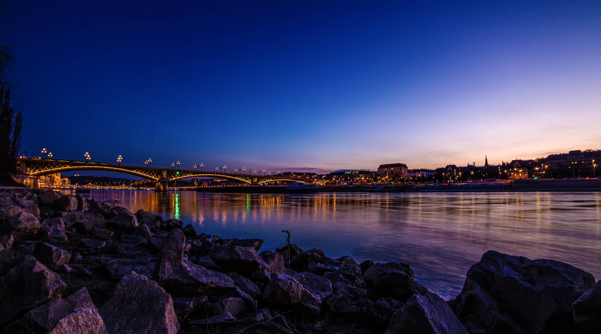 Rocky Riverside With View Of Bridge Background