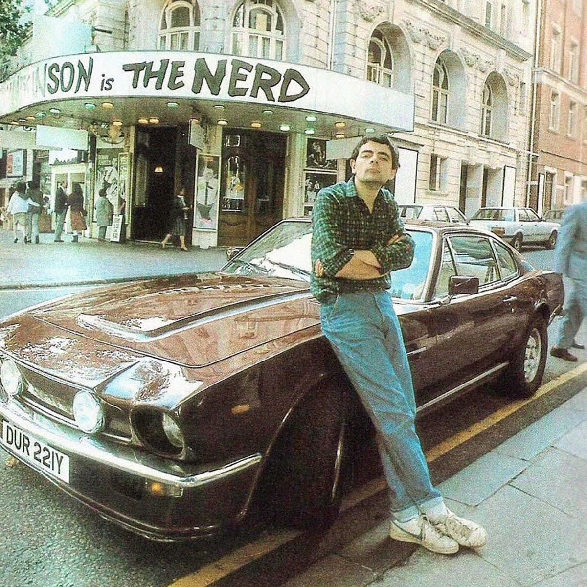 Rowan Atkinson Leaningon Car Outside Theatre Background