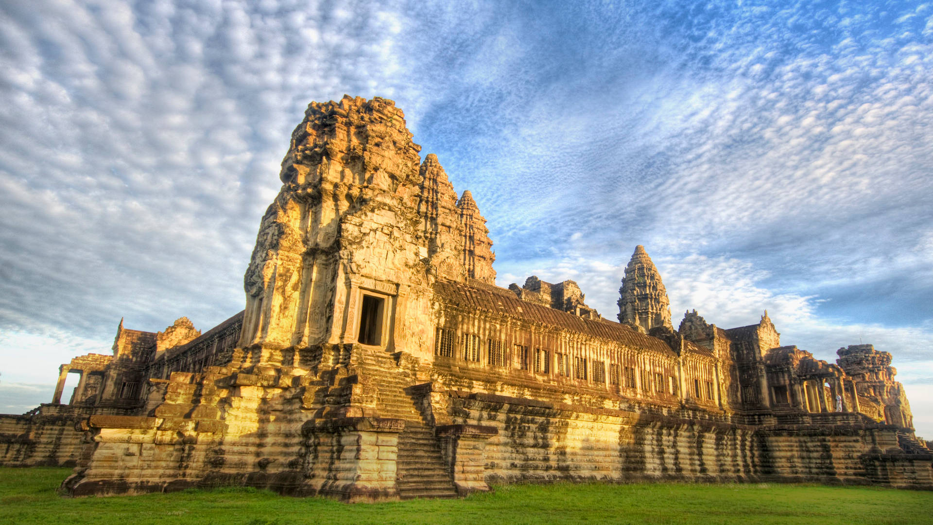 The Majestic Angkor Wat Amidst Verdant Greenery Under A Clear Blue Sky Background