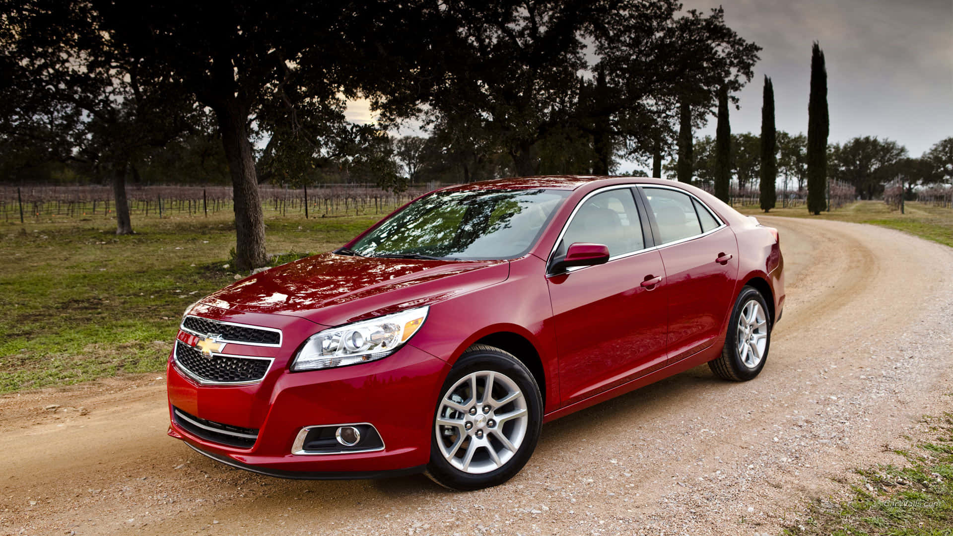 The Red Chevrolet Malibu Is Driving Down A Dirt Road Background