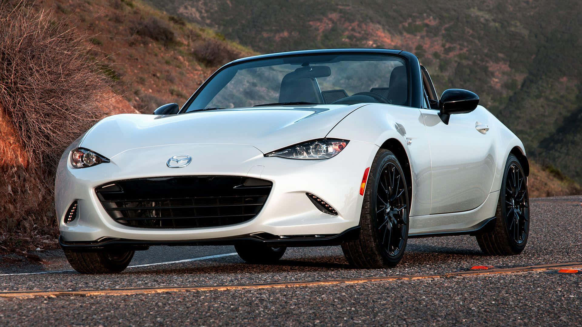 The White Mazda Mx-5 Roadster Is Driving Down A Mountain Road Background