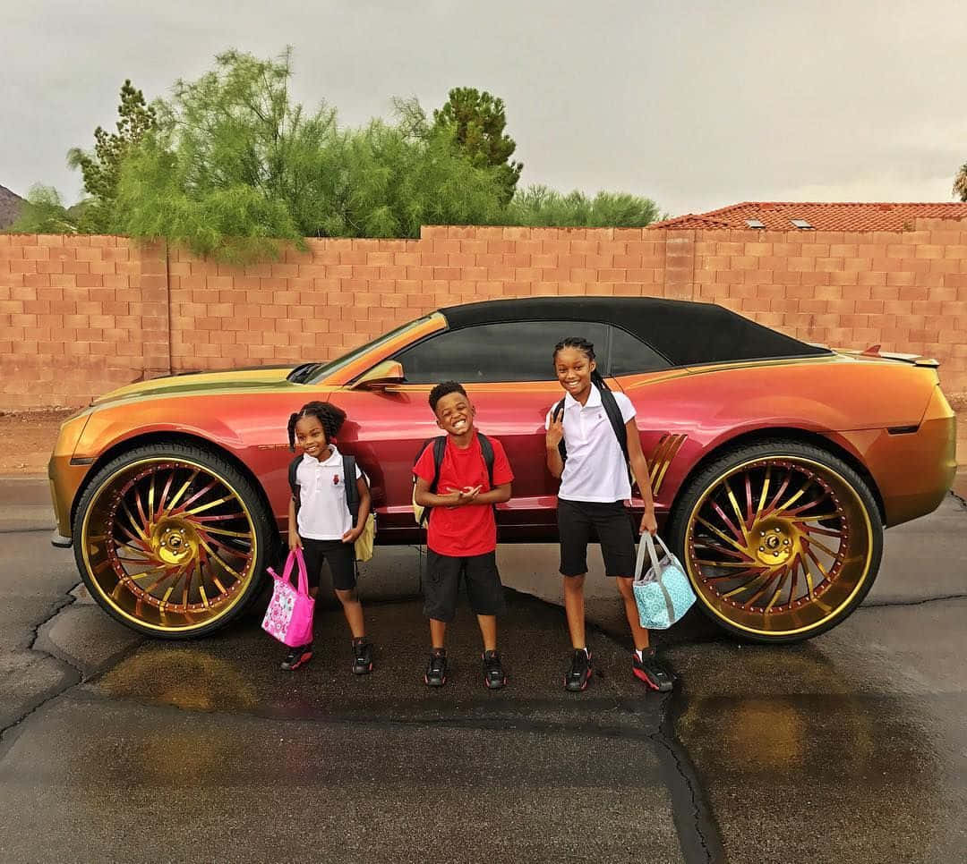 Three Children Standing Next To A Car With Large Wheels Background