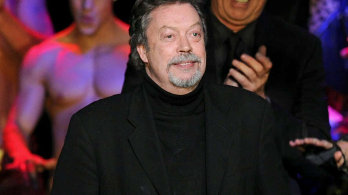 Tim Curry Smiling In A Classic Hollywood Portrait Background