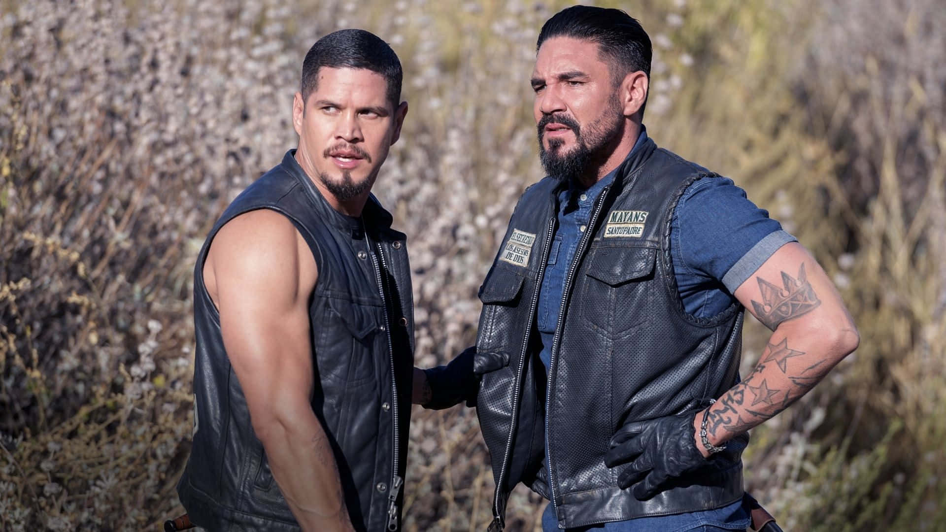 Two Men In Leather Vests Standing In A Field Background