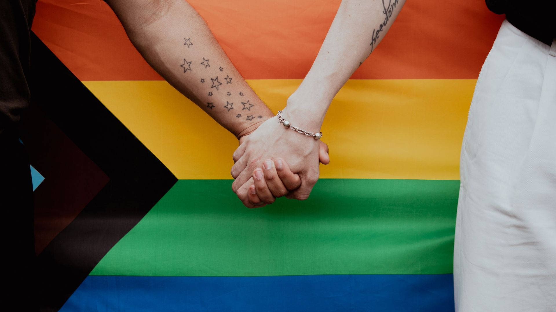 Two People Holding Hands In Front Of A Rainbow Flag Background