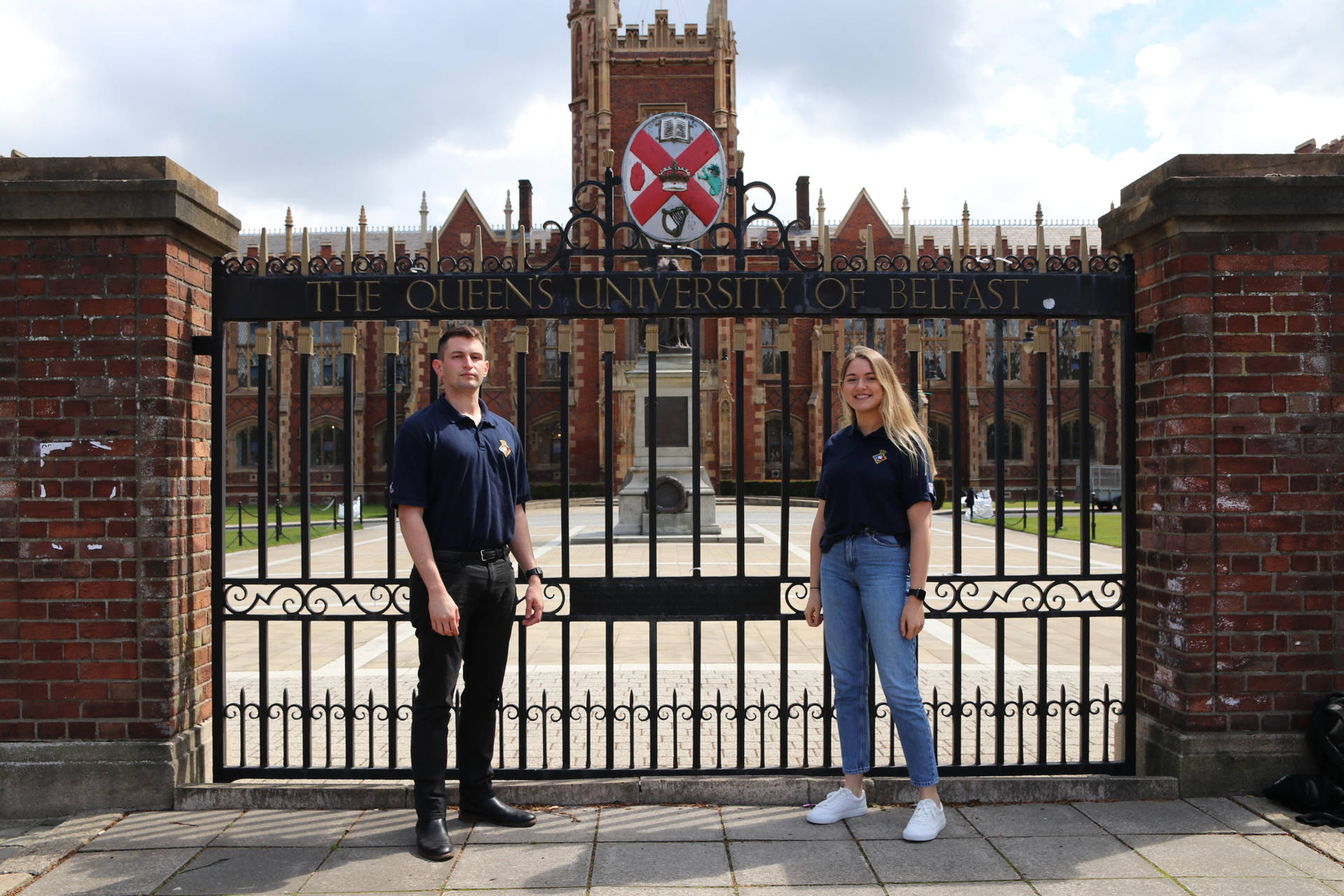 Two People Standing In Front Of A Gated Entrance Background