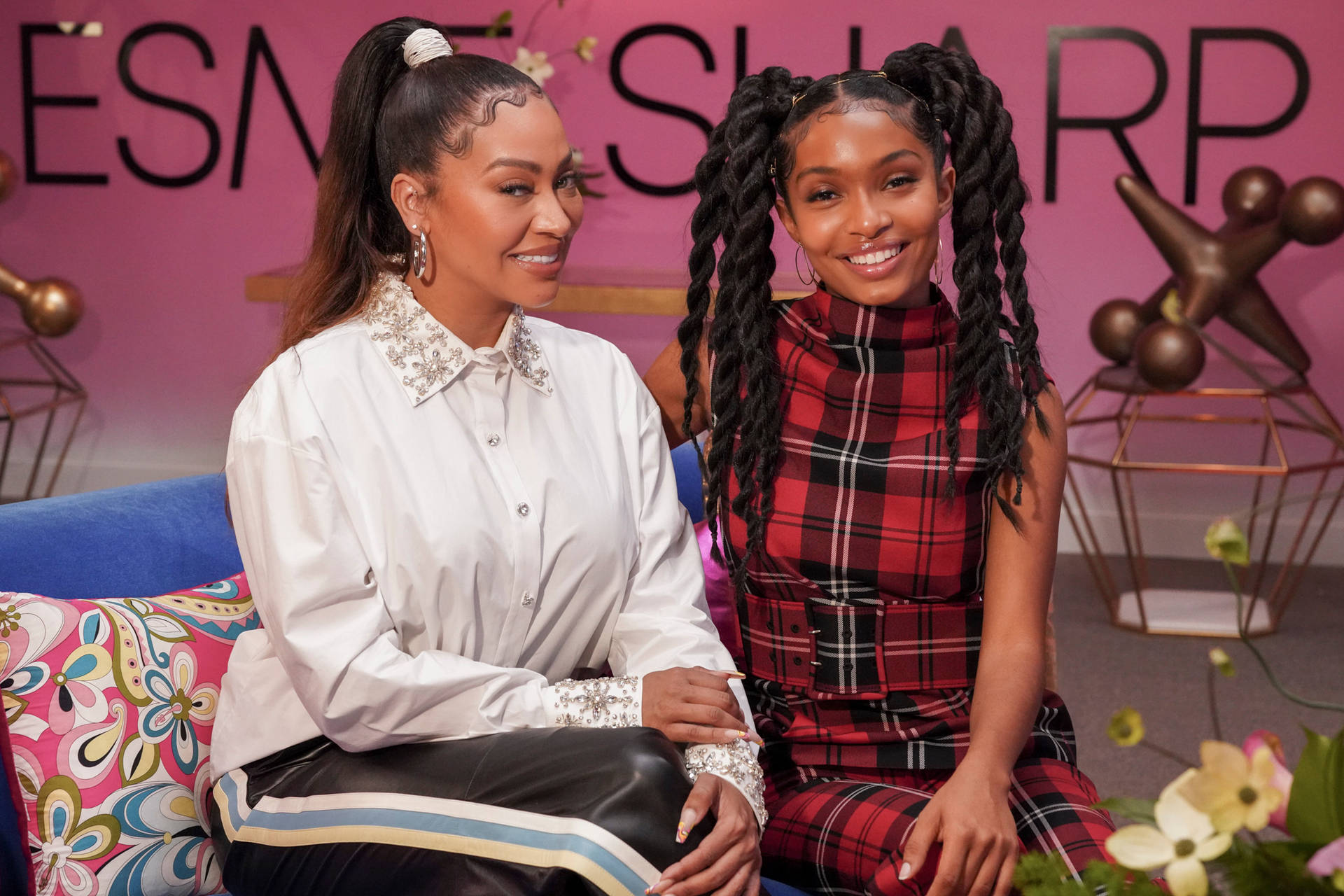 Two Women Sitting On A Couch With A Pink Background Background