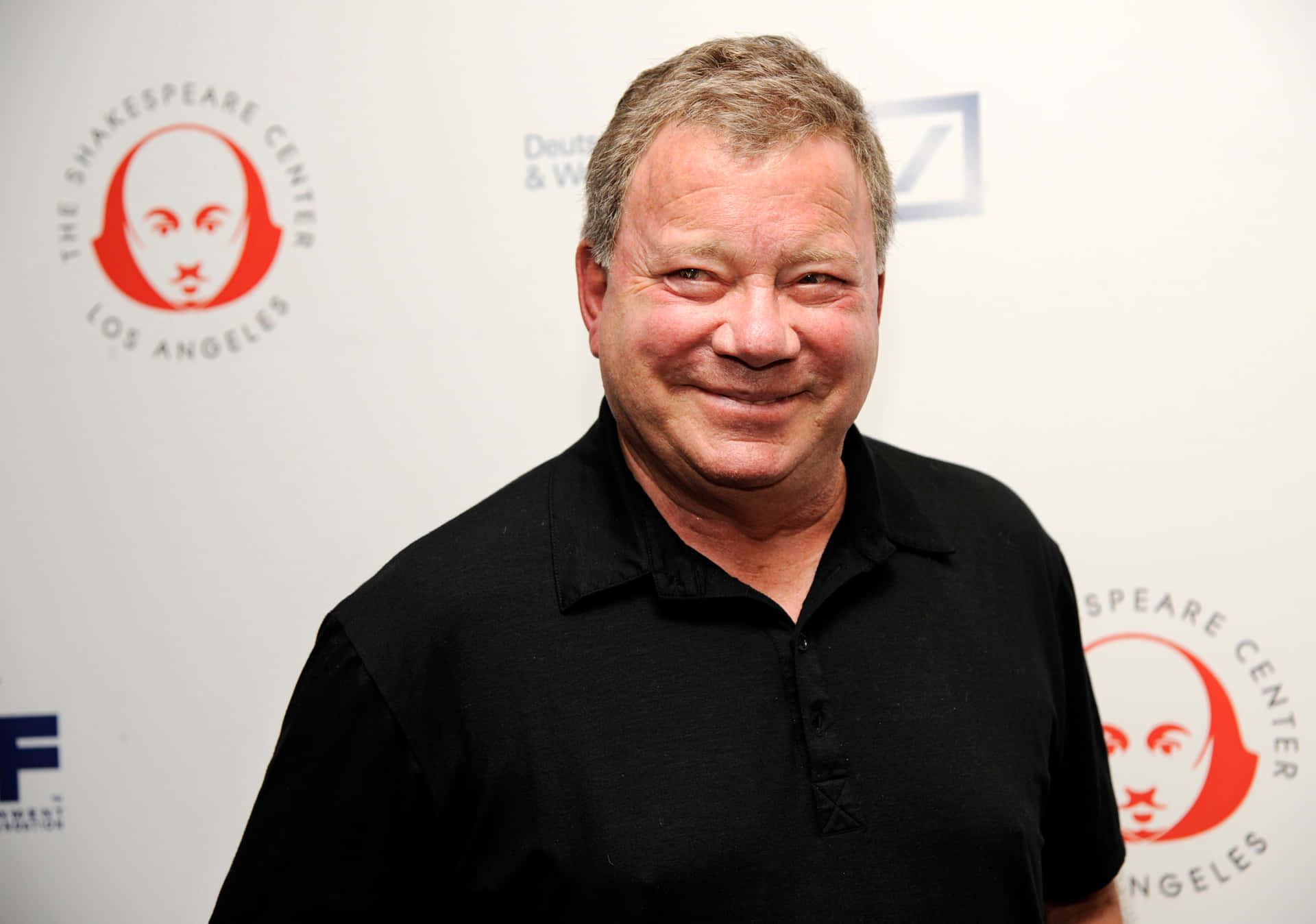 William Shatner Smiling In A Black Suit With A Blue Shirt Background
