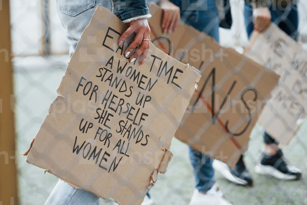 Women Holding Signs That Say Every Time A Woman Stands Up For Herself, She Stands Up For Women Background